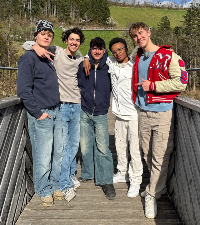 Fünf Jungs stehen auf einer Holzbrücke nebeneinander. Im Hintergrund sind Tannen und grüne Wiesen zu sehen. Die Jungs tragen Jeans und Pullover oder Sweatshirt-Jacken. Sie haben die Arme umeinander gelegt und schauen fröhlich in die Kamera.