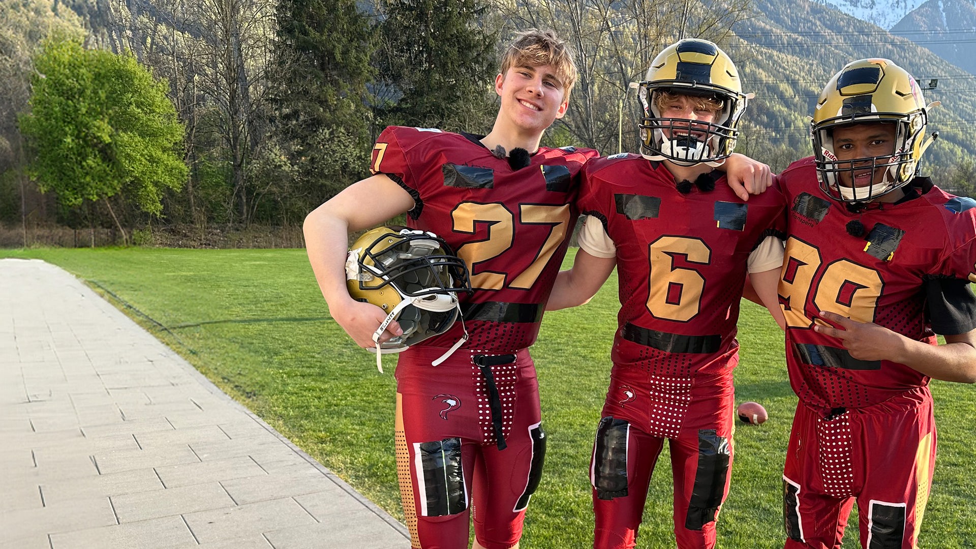 Drei Jungs posten im roten Football-Outfit auf einer grünen Wiese. Im Hintergrund sind Berge zu sehen. Die Jungs tragen die typischen Footballhelme mit Gitter vor dem Gesicht, die Schultern sind ausgepolstert. Luke, Cay und Azad aus der Jungs-WG haben sich gegenseitig die Arme um die Schultern gelegt und lachen in die Kamera.
