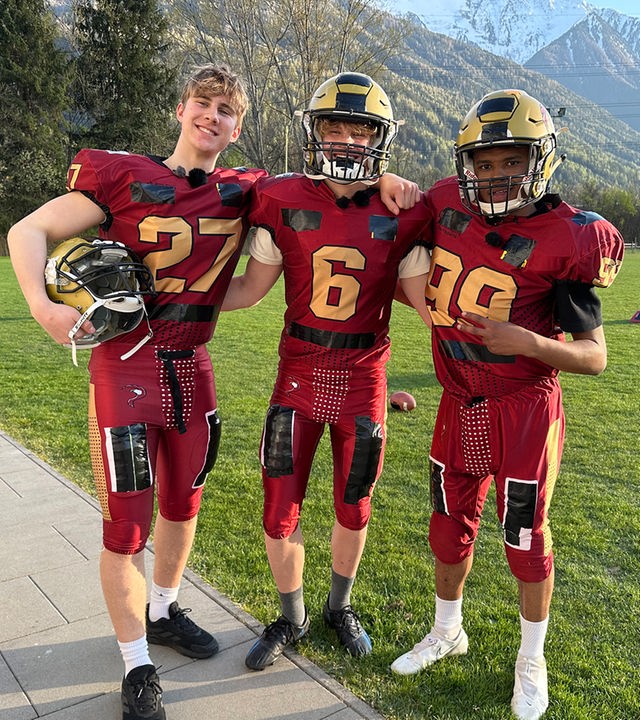Drei Jungs posen im roten Football-Outfit auf einer grünen Wiese. Im Hintergrund sind Berge zu sehen. Die Jungs tragen die typischen Footballhelme mit Gitter vor dem Gesicht, die Schultern sind ausgepolstert. Luke, Cay und Azad aus der Jungs-WG haben sich gegenseitig die Arme um die Schultern gelegt und lachen in die Kamera.