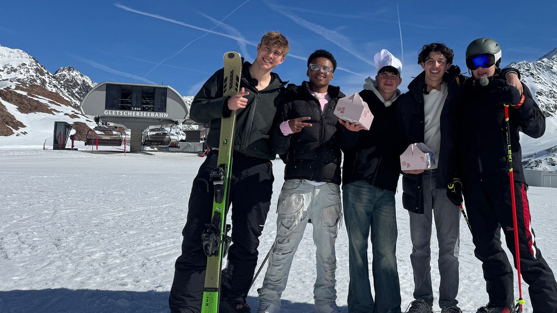 Fünf Jungs stehen nebeneinander auf einer schneebedeckten Skipiste. Im Hintergrund ist ein Sessellift zu sehen. Die Jungs jeweils ganz rechts und links außen tragen Skiklamotten und halten Skier oder Stöcke in den Händen. Es sind Luke und Cay, die heute einen Freestyle Kurs geschenkt bekommen haben. Die drei Jungs in der Mitte - Azad, Nick und Lorian - tragen Jeans und halten rosafarbene Pakete in den Händen. Nick trägt außerdem eine Kochmütze auf dem Kopf. Die drei waren beim Praktikum in der höchsten Konditorei Österreichs.