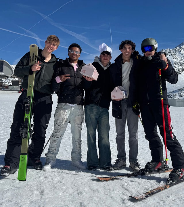 Fünf Jungs stehen nebeneinander auf einer schneebedeckten Skipiste. Im Hintergrund ist ein Sessellift zu sehen. Die Jungs jeweils ganz rechts und links außen tragen Skiklamotten und halten Skier oder Stöcke in den Händen. Es sind Luke und Cay, die heute einen Freestyle Kurs geschenkt bekommen haben. Die drei Jungs in der Mitte - Azad, Nick und Lorian - tragen Jeans und halten rosafarbene Pakete in den Händen. Nick trägt außerdem eine Kochmütze auf dem Kopf. Die drei waren beim Praktikum in der höchsten Konditorei Österreichs.