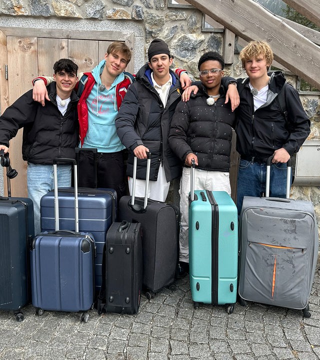 Fünf Jungs stehen nebeneinander vor eine Hauswand aus Holz und Steinen. Sie haben die Arme umeinander gelegt und halten gleichzeitig mit einer Hand den Griff eines Rollkoffers. Für Nick, Luke, Lorian, Azad und Cay ist die Zeit in der Jungs-WG zu Ende.
