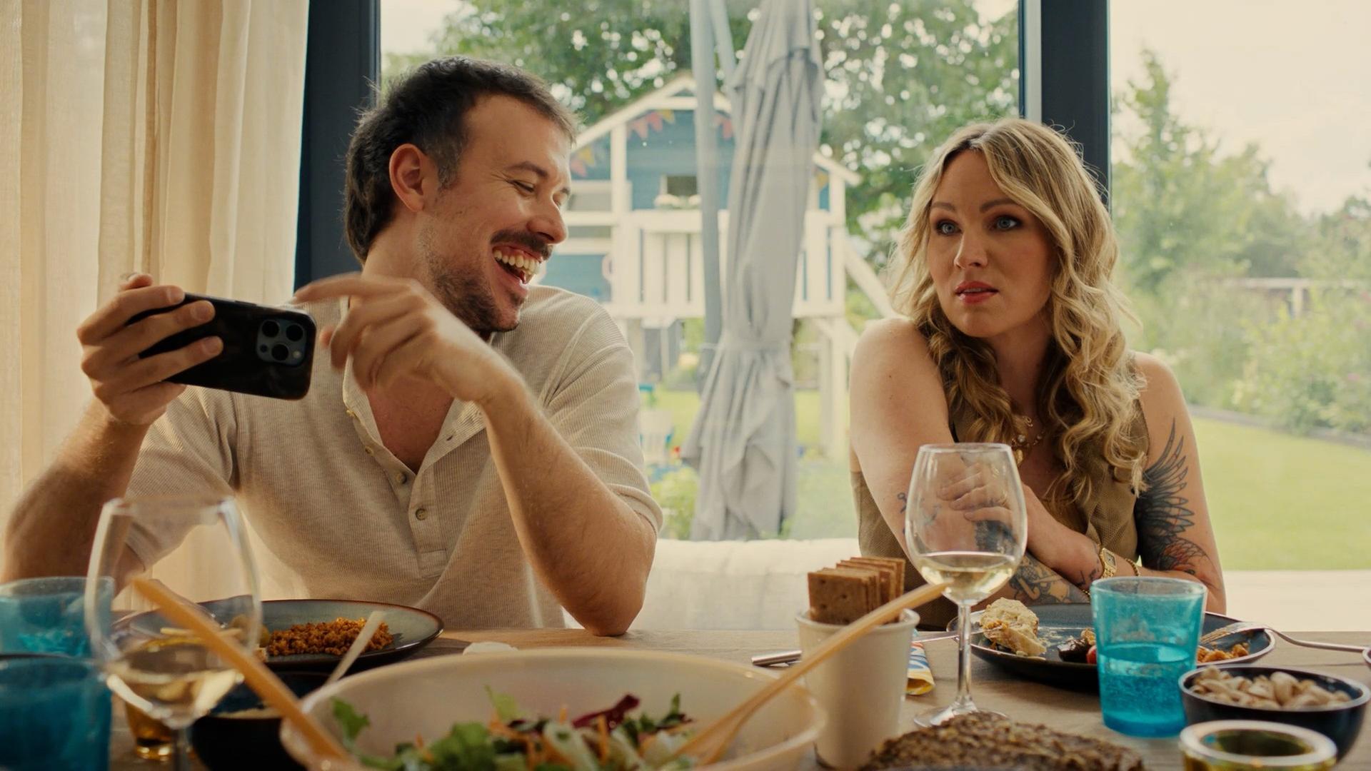 "The other gAIrl – Jurassic Spark": Patrick (Fred Costea) und Christin (Evelyn Weigert) sitzen am gedeckten Tisch. Patrick lacht und hält ein Smartphone in den Händen.