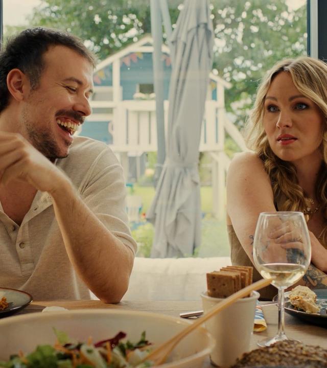 "The other gAIrl – Jurassic Spark": Patrick (Fred Costea) und Christin (Evelyn Weigert) sitzen am gedeckten Tisch. Patrick lacht und hält ein Smartphone in den Händen.