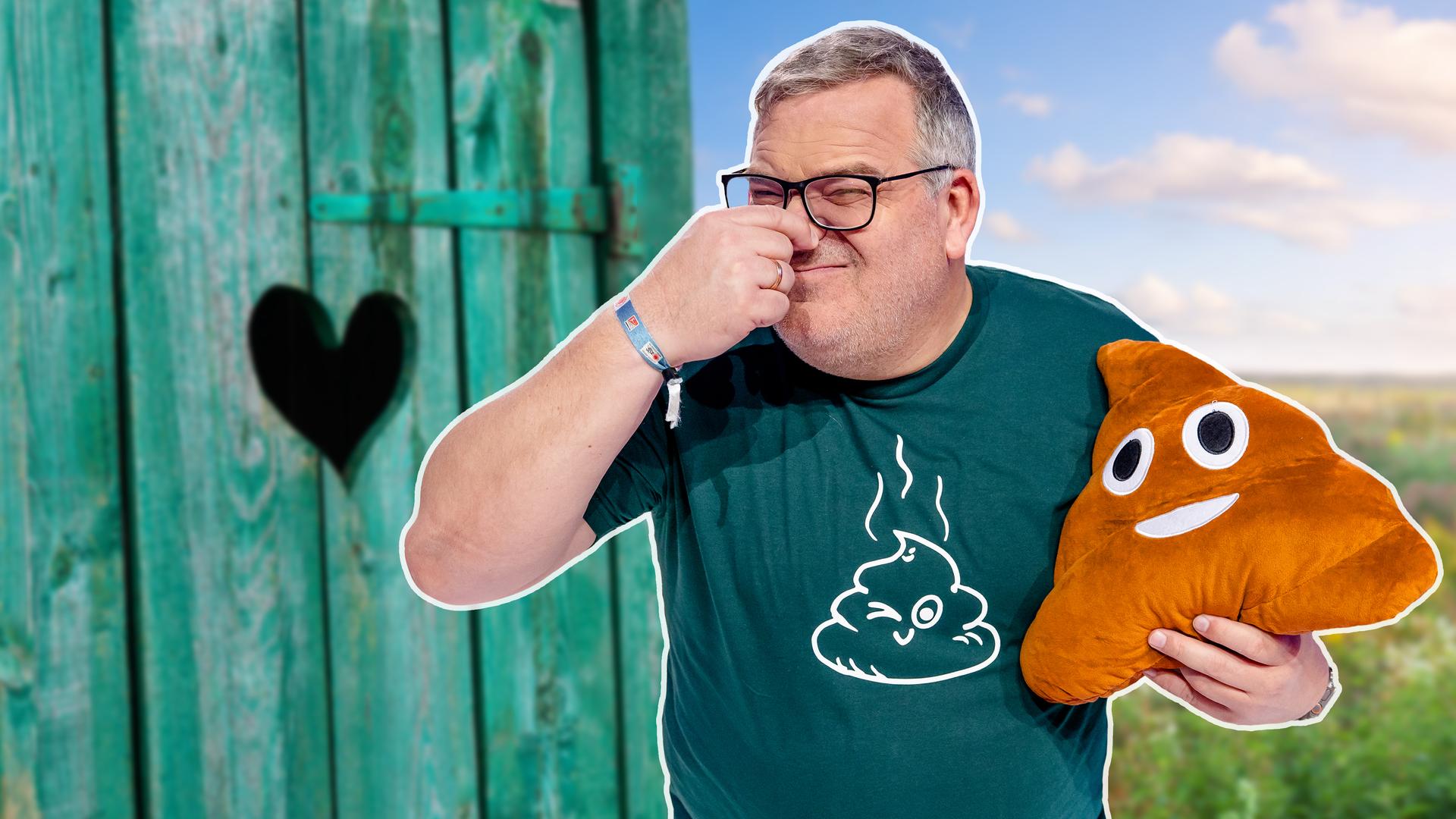 Elton steht vor einem Klo-Häuschen, hält sich die Nase zu und trägt ein Kack-Emoj-Kissen unter dem Arm. 
