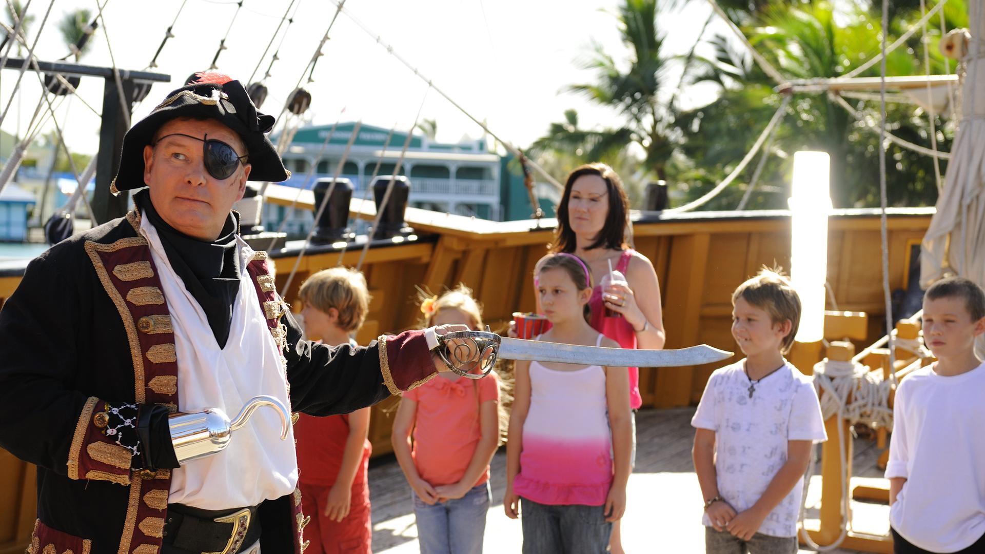 Don steht auf einem Piratenschiff und ist als Piraten-Käpt'n verkleidet: Er trägt Augenklappe und Piratenhut und hält mit ausgestrecktem Arm einen Säbel vor sich. Im Hintergrund stehen eine Gruppe Kinder und eine Frau, die ihn beobachten.
