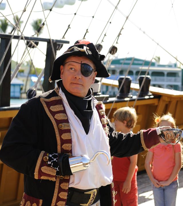 Don steht auf einem Piratenschiff und ist als Piraten-Käpt'n verkleidet: Er trägt Augenklappe und Piratenhut und hält mit ausgestrecktem Arm einen Säbel vor sich. Im Hintergrund stehen eine Gruppe Kinder und eine Frau, die ihn beobachten.
