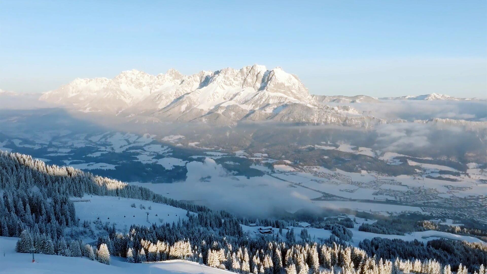 Das Bild zeigt eine beeindruckende winterliche Landschaft im Kaisergebirge in Tirol. Die Szenerie ist geprägt von hohen, schneebedeckten Bergen, die im Hintergrund thronen, während im Vordergrund dichte Nadelwälder zu sehen sind, die ebenfalls mit Schnee bedeckt sind. Die Bäume sind teilweise sonnendurchflutet und erscheinen heller, während der Rest der Landschaft schattiger ist.   Der Himmel ist klar und blau, was die Kälte und Frische des Morgens unterstreicht. Im Tal erstreckt sich eine weite, schneebedeckte Landschaft, die von einzelnen Nebelschwaden durchzogen ist. Hier sind auch sporadisch Wohngebäude und Straßen sichtbar, die sich durch die winterliche Landschaft schlängeln.   Insgesamt vermittelt das Bild eine ruhige und friedliche Winteratmosphäre, die die natürliche Schönheit der Umgebung hervorhebt. Es handelt sich um eine Szene, die typisch für die Region St. Johann in Tirol ist und die sportlichen Abenteuer sowie das Leben der Menschen in dieser einzigartigen Gebirgslandschaft präsentiert.