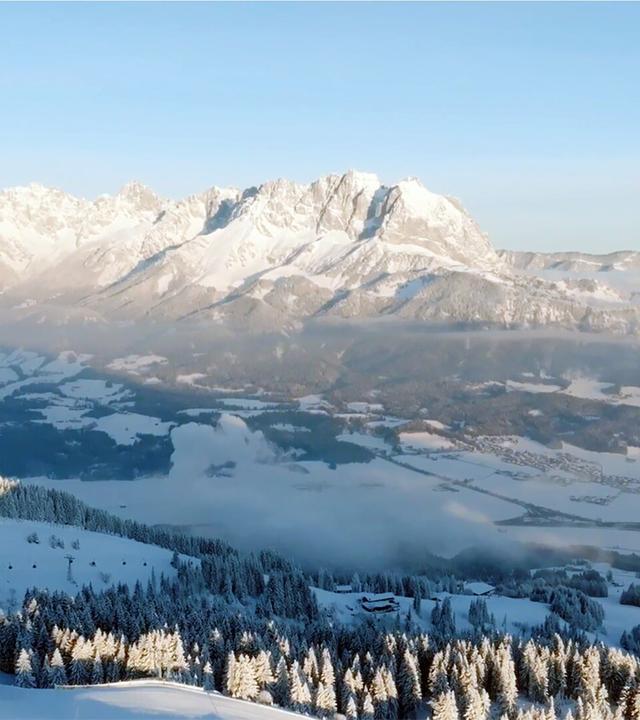 Das Bild zeigt eine beeindruckende winterliche Landschaft im Kaisergebirge in Tirol. Die Szenerie ist geprägt von hohen, schneebedeckten Bergen, die im Hintergrund thronen, während im Vordergrund dichte Nadelwälder zu sehen sind, die ebenfalls mit Schnee bedeckt sind. Die Bäume sind teilweise sonnendurchflutet und erscheinen heller, während der Rest der Landschaft schattiger ist.   Der Himmel ist klar und blau, was die Kälte und Frische des Morgens unterstreicht. Im Tal erstreckt sich eine weite, schneebedeckte Landschaft, die von einzelnen Nebelschwaden durchzogen ist. Hier sind auch sporadisch Wohngebäude und Straßen sichtbar, die sich durch die winterliche Landschaft schlängeln.   Insgesamt vermittelt das Bild eine ruhige und friedliche Winteratmosphäre, die die natürliche Schönheit der Umgebung hervorhebt. Es handelt sich um eine Szene, die typisch für die Region St. Johann in Tirol ist und die sportlichen Abenteuer sowie das Leben der Menschen in dieser einzigartigen Gebirgslandschaft präsentiert.