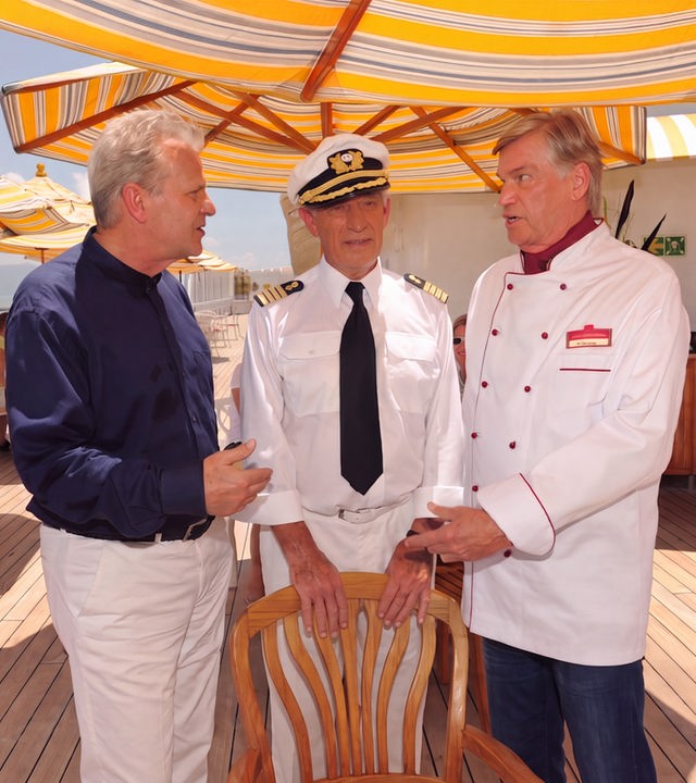 "Das Traumschiff: Kambodscha": Der Spitzenkoch Philipp Mejée (Peter Sattmann) diskutiert mit dem Schiffsmaître Dieter Walde (Jochen Busse) an Deck. Kapitän Paulsen (Siegfried Rauch) steht zwischen beiden und ist sichtlich genervt von den Streithähnen.