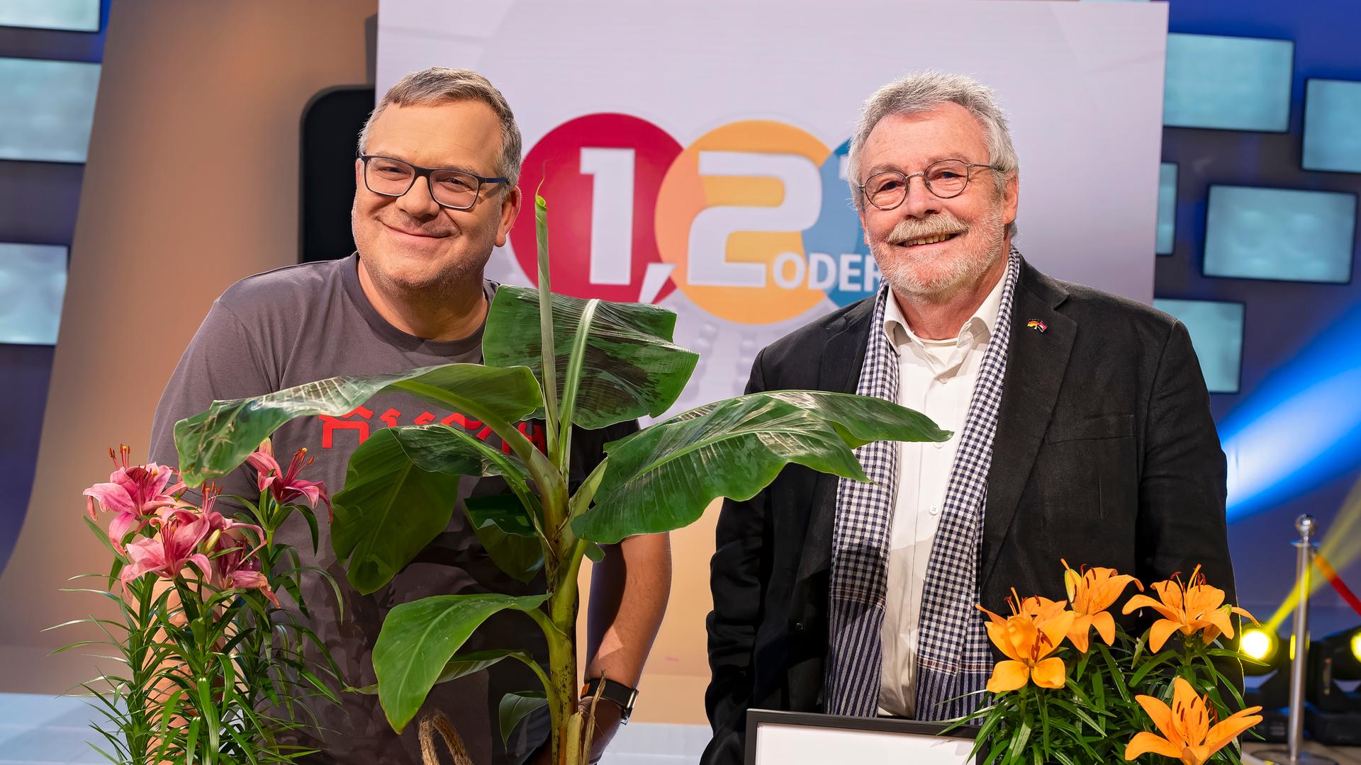 Elton und sein Gast Prof. Dr. Hans Leisen stehen neben einem Tisch mit Gegenständen, die typisch für Kambodscha sind. Auf dem Tisch stehen zum Beispiel Pflanzen, Bananen, Getränkedosen und die Nationalflagge Kambodschas. Elton hält eine Mango in der Hand.