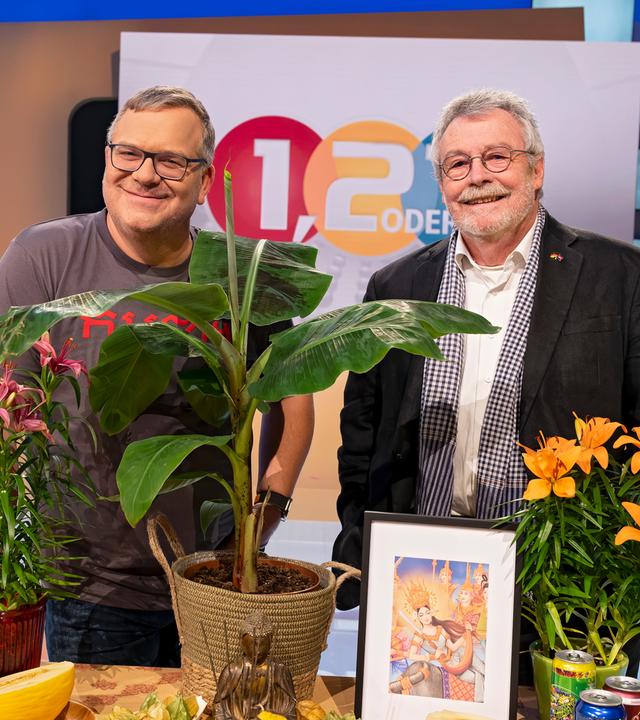 Elton und sein Gast Prof. Dr. Hans Leisen stehen neben einem Tisch mit Gegenständen, die typisch für Kambodscha sind. Auf dem Tisch stehen zum Beispiel Pflanzen, Bananen, Getränkedosen und die Nationalflagge Kambodschas. Elton hält eine Mango in der Hand.
