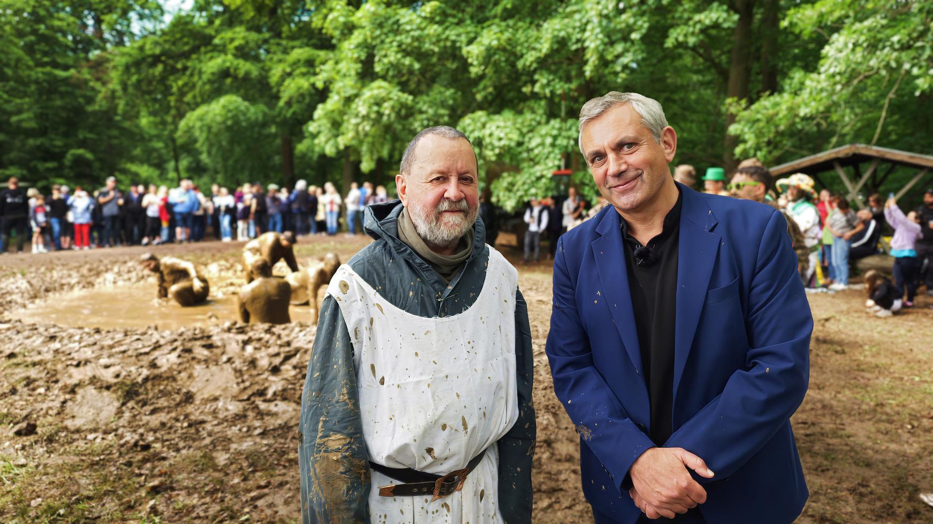Vladimir Kaminer im blauen Anzug und eine Mann in einem weißen, schmutzigen Umhang, stehen lächelnd vor einer Gruppe von Menschen, die im Hintergrund zusehen. Einige Personen im Hintergrund spielen im Matsch. Im Freien, Bäume und Grün zu sehen.