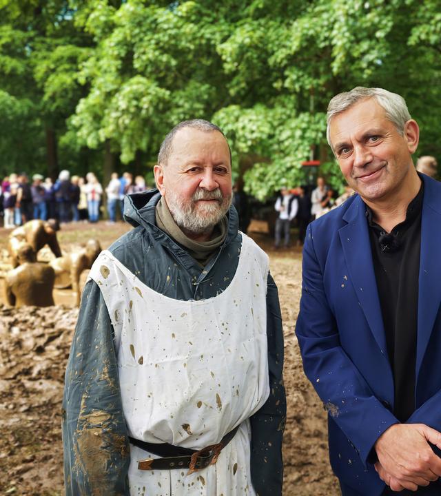 Vladimir Kaminer im blauen Anzug und eine Mann in einem weißen, schmutzigen Umhang, stehen lächelnd vor einer Gruppe von Menschen, die im Hintergrund zusehen. Einige Personen im Hintergrund spielen im Matsch. Im Freien, Bäume und Grün zu sehen.