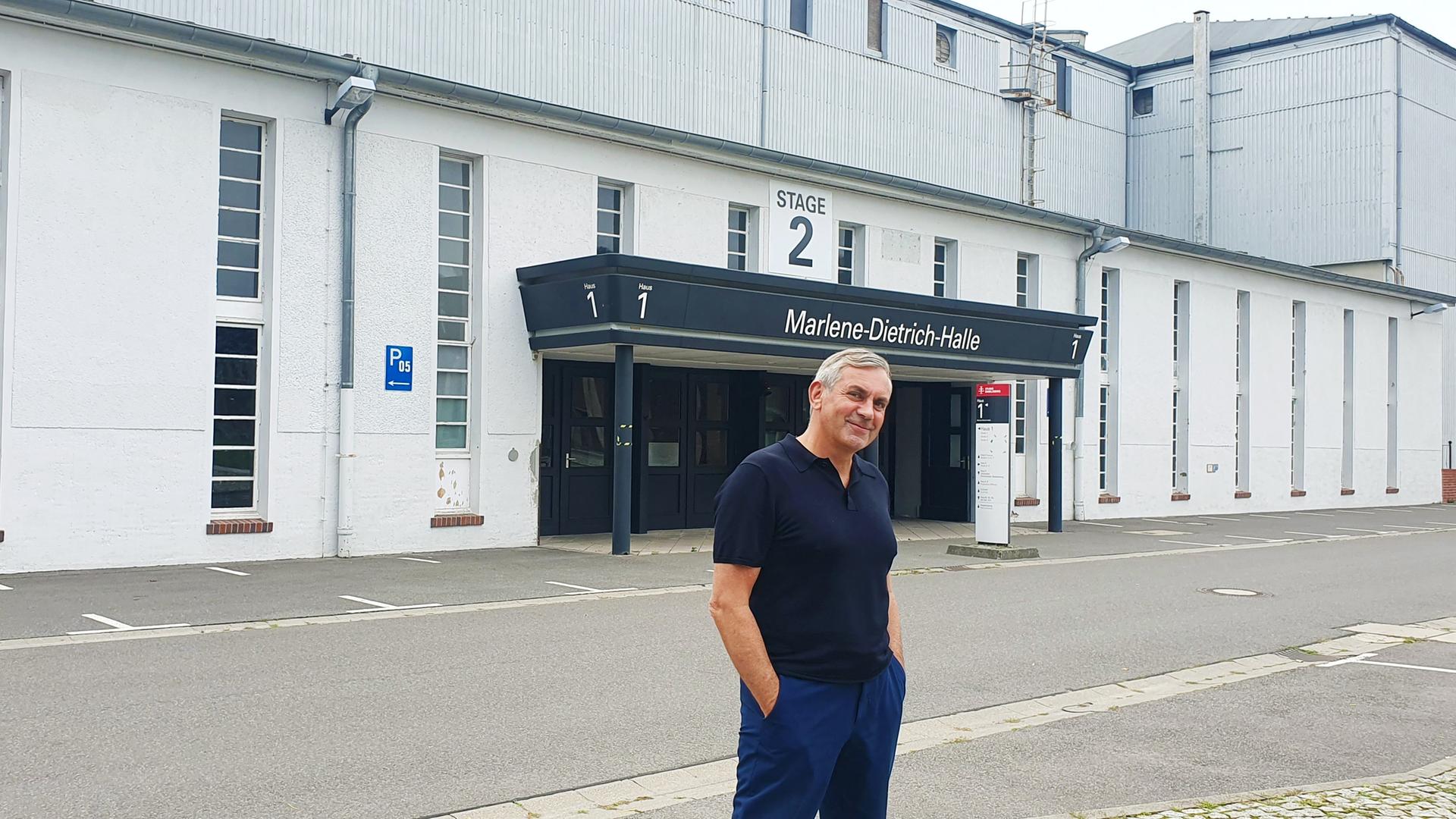 Wladimir Kaminer steht lässig mit blauem Poloshirt und blauer Hose, Hände in den Hosentaschen vor einer Hallenfront