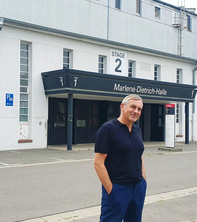Wladimir Kaminer steht lässig mit blauem Poloshirt und blauer Hose, Hände in den Hosentaschen vor einer Hallenfront