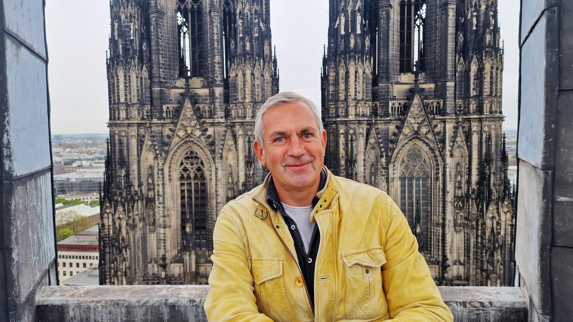Ein Mann in gelber Lederjacke lehnt an einer steinernen Brüstung vor den Türmen des Kölner Doms; Blick über die Stadt Köln im Hintergrund. 