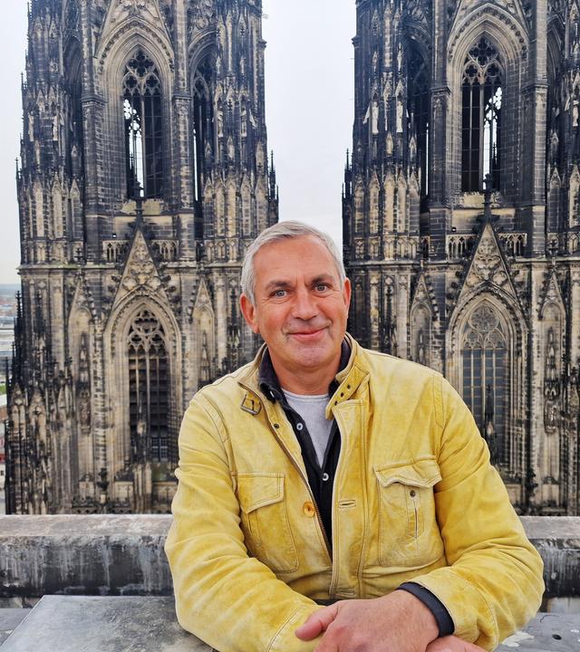 Ein Mann in gelber Lederjacke lehnt an einer steinernen Brüstung vor den Türmen des Kölner Doms; Blick über die Stadt Köln im Hintergrund. 