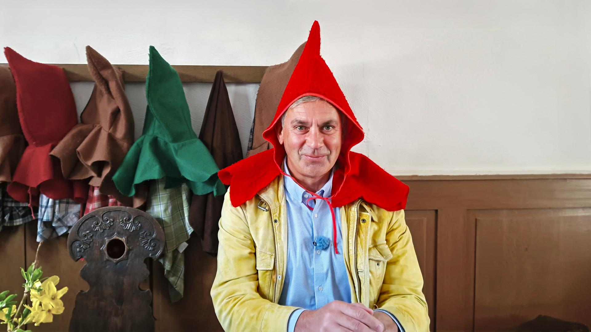 Wladimir Kaminer mit rotem Spitzhut und beiger Jacke an einem Tisch; im Hintergrund hängen bunte Hüte an der Wand -- Szene aus Kaminer Inside: Deutsche Märchenstraße.