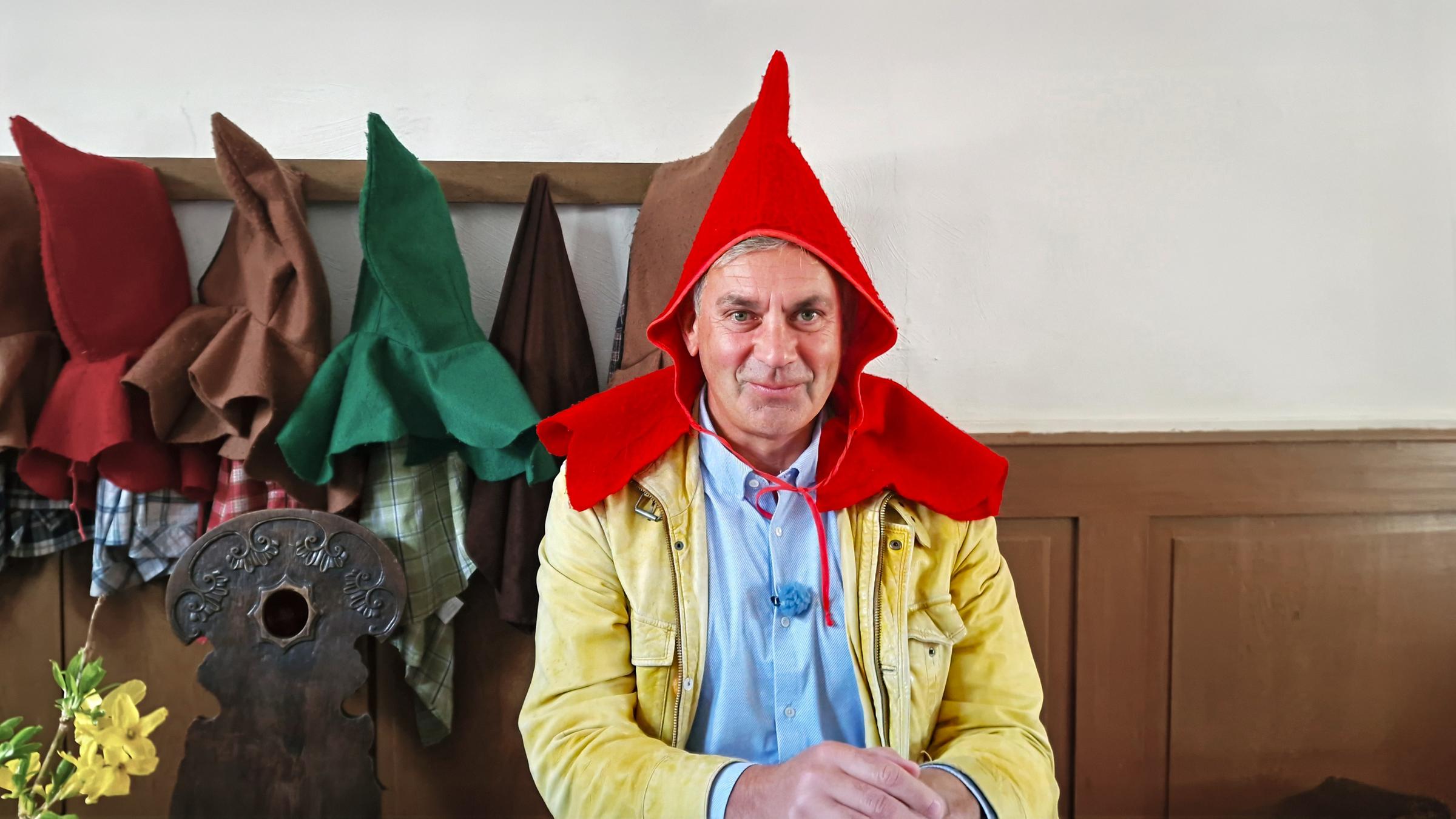 Wladimir Kaminer mit rotem Spitzhut und beiger Jacke an einem Tisch; im Hintergrund hängen bunte Hüte an der Wand -- Szene aus Kaminer Inside: Deutsche Märchenstraße.