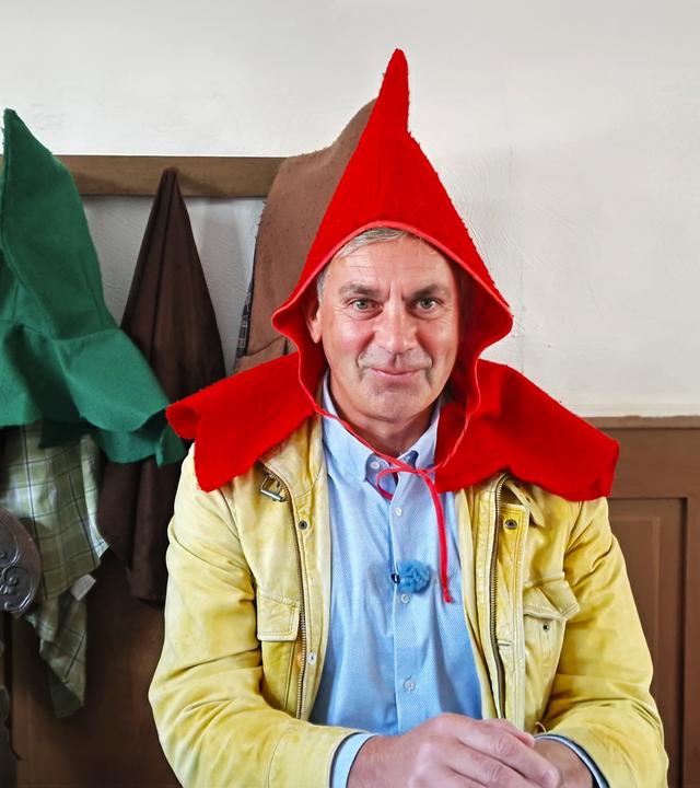 Wladimir Kaminer mit rotem Spitzhut und beiger Jacke an einem Tisch; im Hintergrund hängen bunte Hüte an der Wand -- Szene aus Kaminer Inside: Deutsche Märchenstraße.