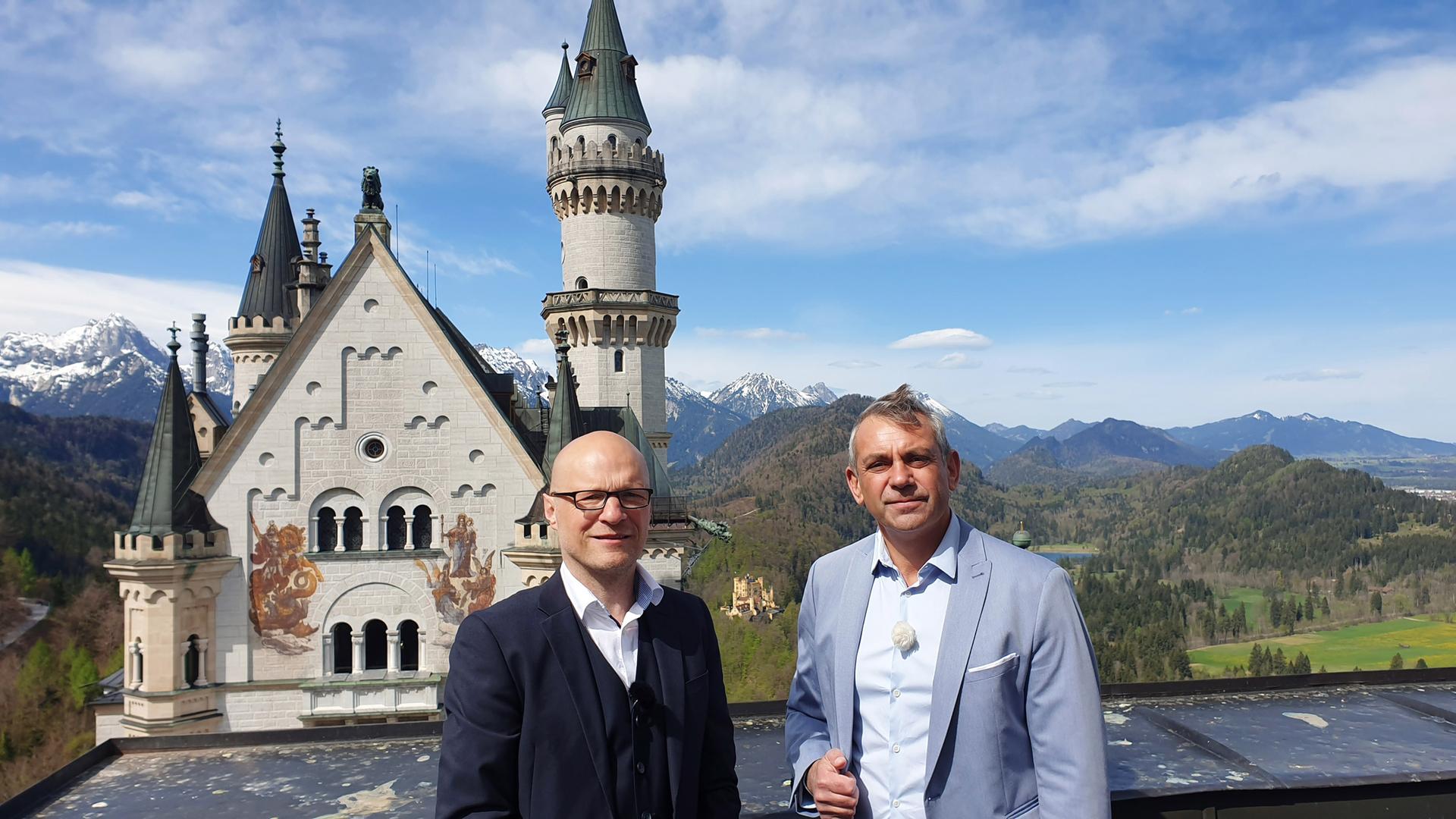 Kaminer und der Schlossherr von Neuschwanstein stehen nebeneinander auf einer Terrasse von Neuschwanstein, hinter ihnen Berge und Wälder
