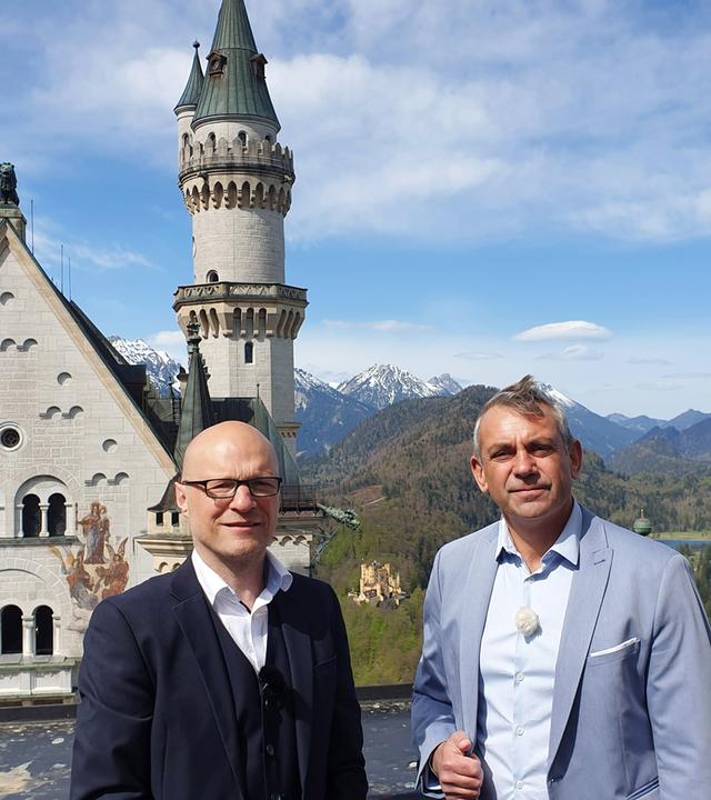 Kaminer und der Schlossherr von Neuschwanstein stehen nebeneinander auf einer Terrasse von Neuschwanstein, hinter ihnen Berge und Wälder