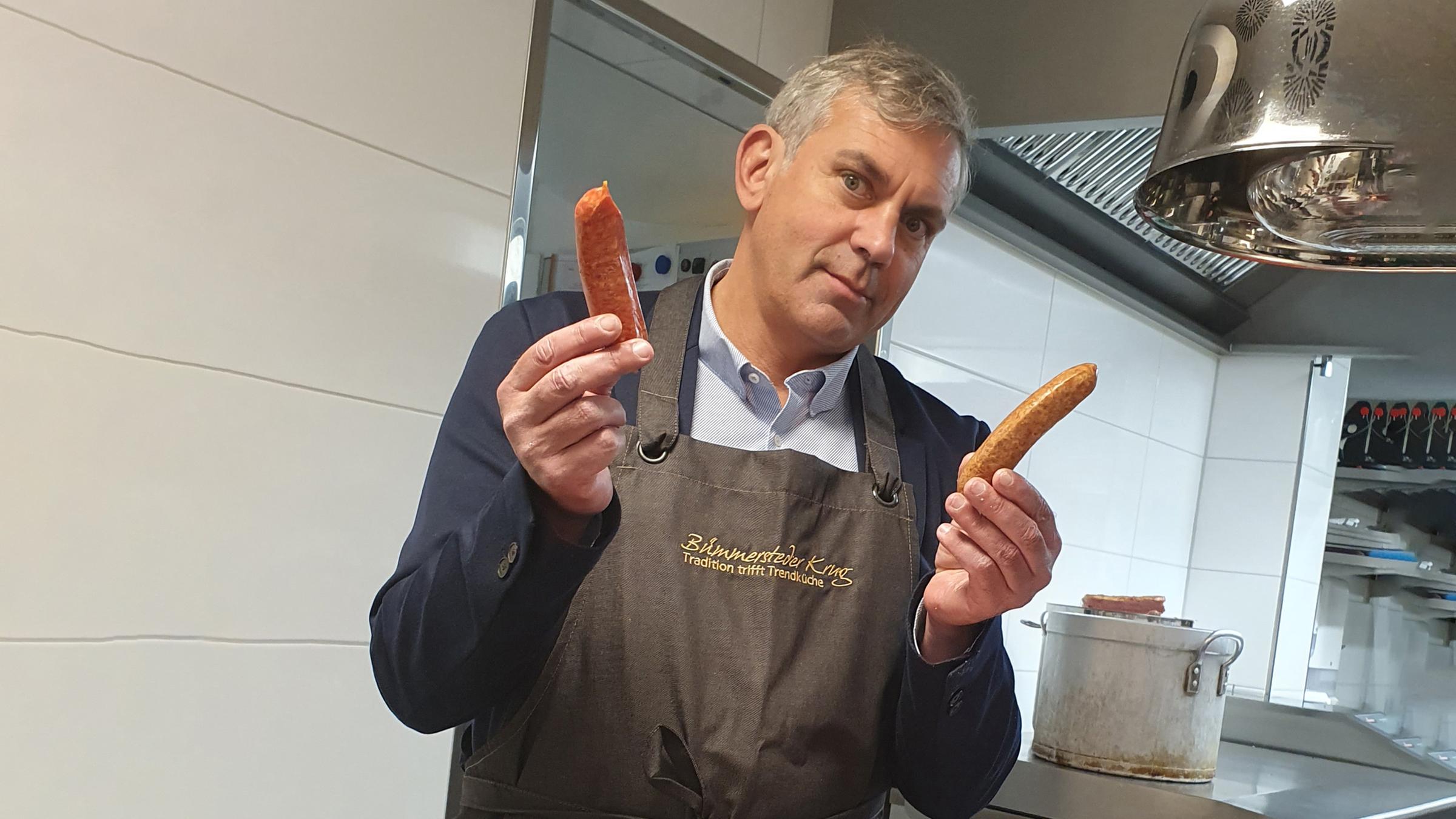 Wladimir Kaminer hält zwei Würstchen in der Hand. Vor ihm eine Schale mit Wurstwaren