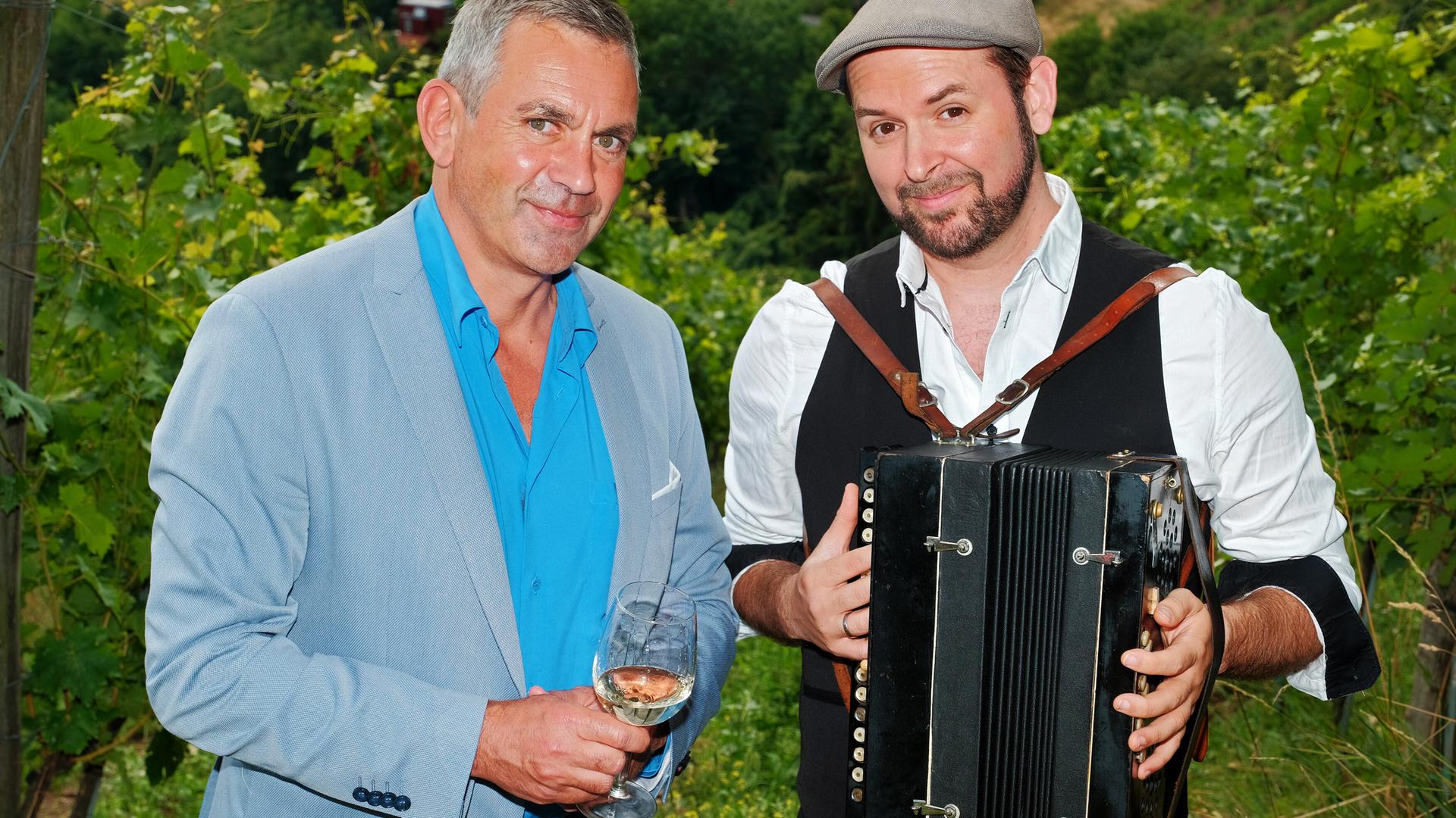 Zwei Männer stehen in einem Weinberg, einer hält ein Glas Wein, der andere ein Akkordeon.