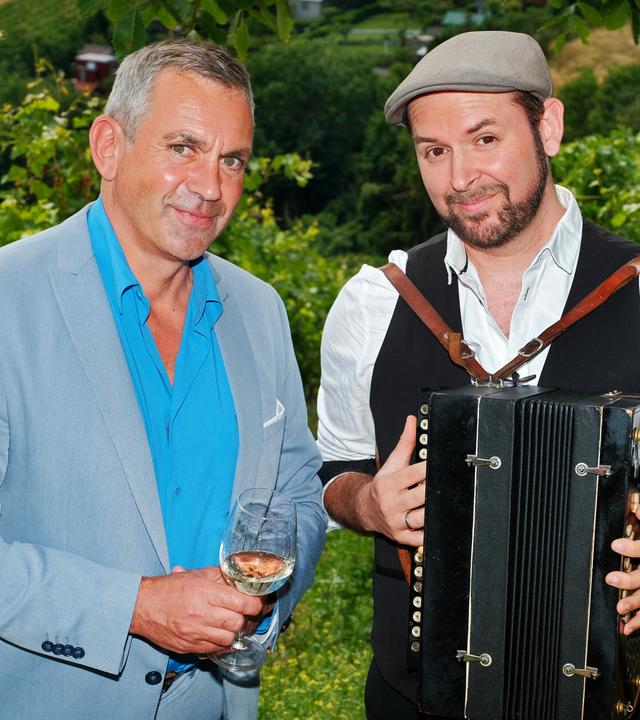 Zwei Männer stehen in einem Weinberg, einer hält ein Glas Wein, der andere ein Akkordeon.