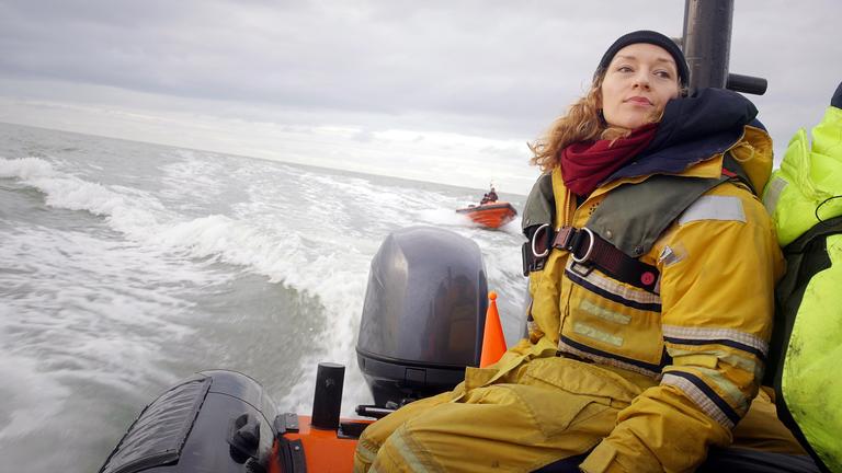 Franziska Saalmann von Greenpeace in gelbem Overall und olivgrüner Schwimmweste fährt im Schlauchboot auf grauer See. Im Hintergrund sieht man ein rotes Boot mit 2 Personen.
