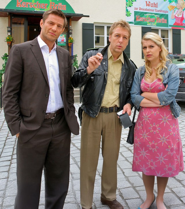 "Die Rosenheim-Cops - Kampf um die Krone": Vor dem Brauereigasthof "Kerscher Bräu" stehen Kommissar Hansen (Igor Jeftic), Polizist Mohr (Max Müller) und Christin Lange (Sarah Thonig). Sie schauen in dieselbe Richtung. Mohr hält einen Notizblock und einen Stift in den Händen.