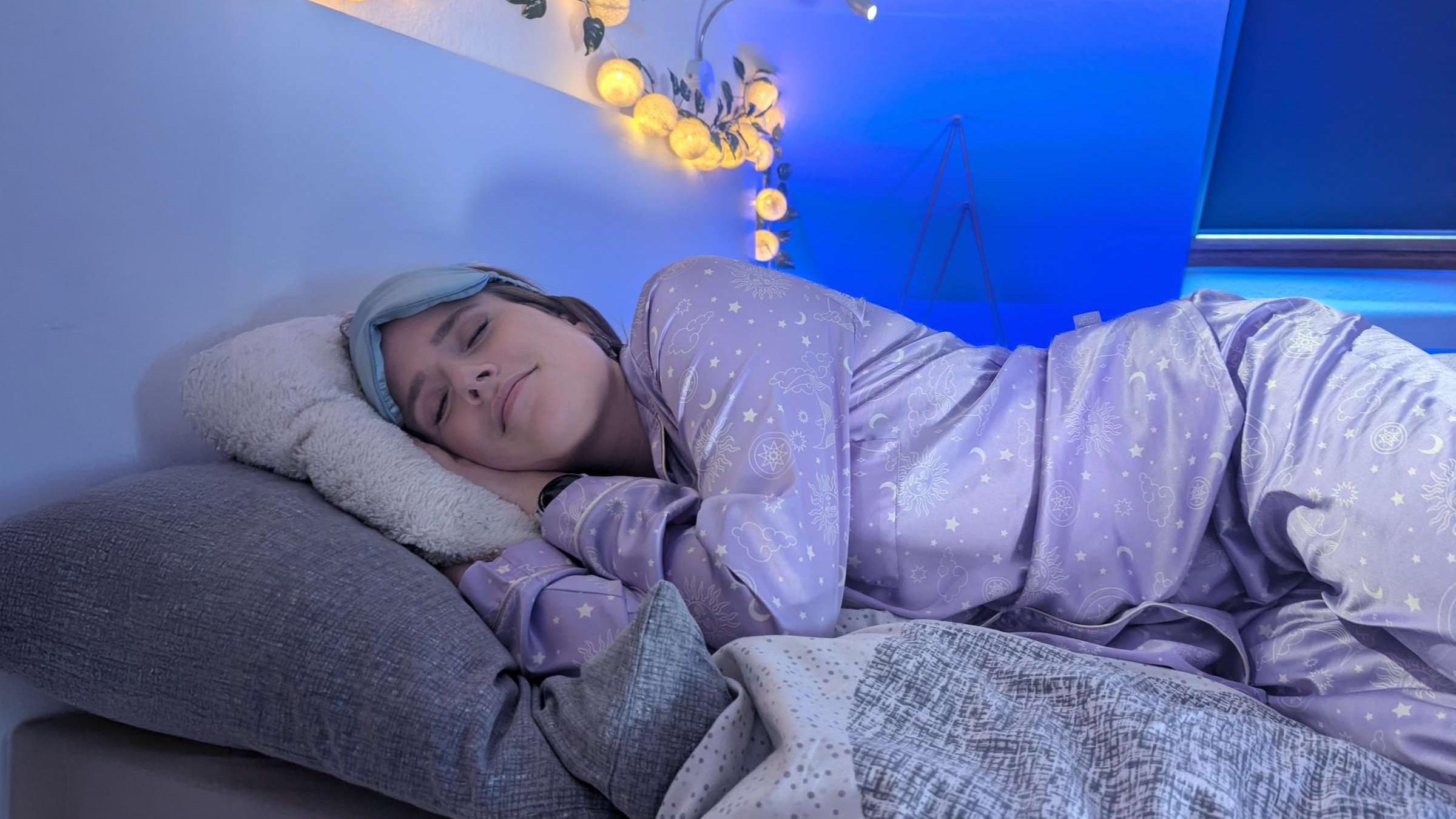 Comedian Saskia Fröhlich liegt in einem Bett auf der Seite und hat ihren Kopf auf ihre Hände gelegt. Ihre Augen sind geschlossen, sie trägt eine Schlafmaske und sie lächelt leicht. Der Raum ist in blaues Licht getaucht, im Hintergrund hängt eine Lichterkette.