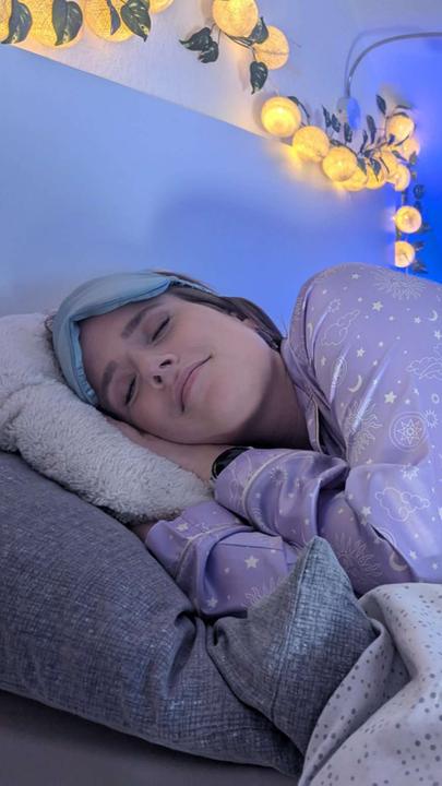 Comedian Saskia Fröhlich liegt in einem Bett auf der Seite und hat ihren Kopf auf ihre Hände gelegt. Ihre Augen sind geschlossen, sie trägt eine Schlafmaske und sie lächelt leicht. Der Raum ist in blaues Licht getaucht, im Hintergrund hängt eine Lichterkette.