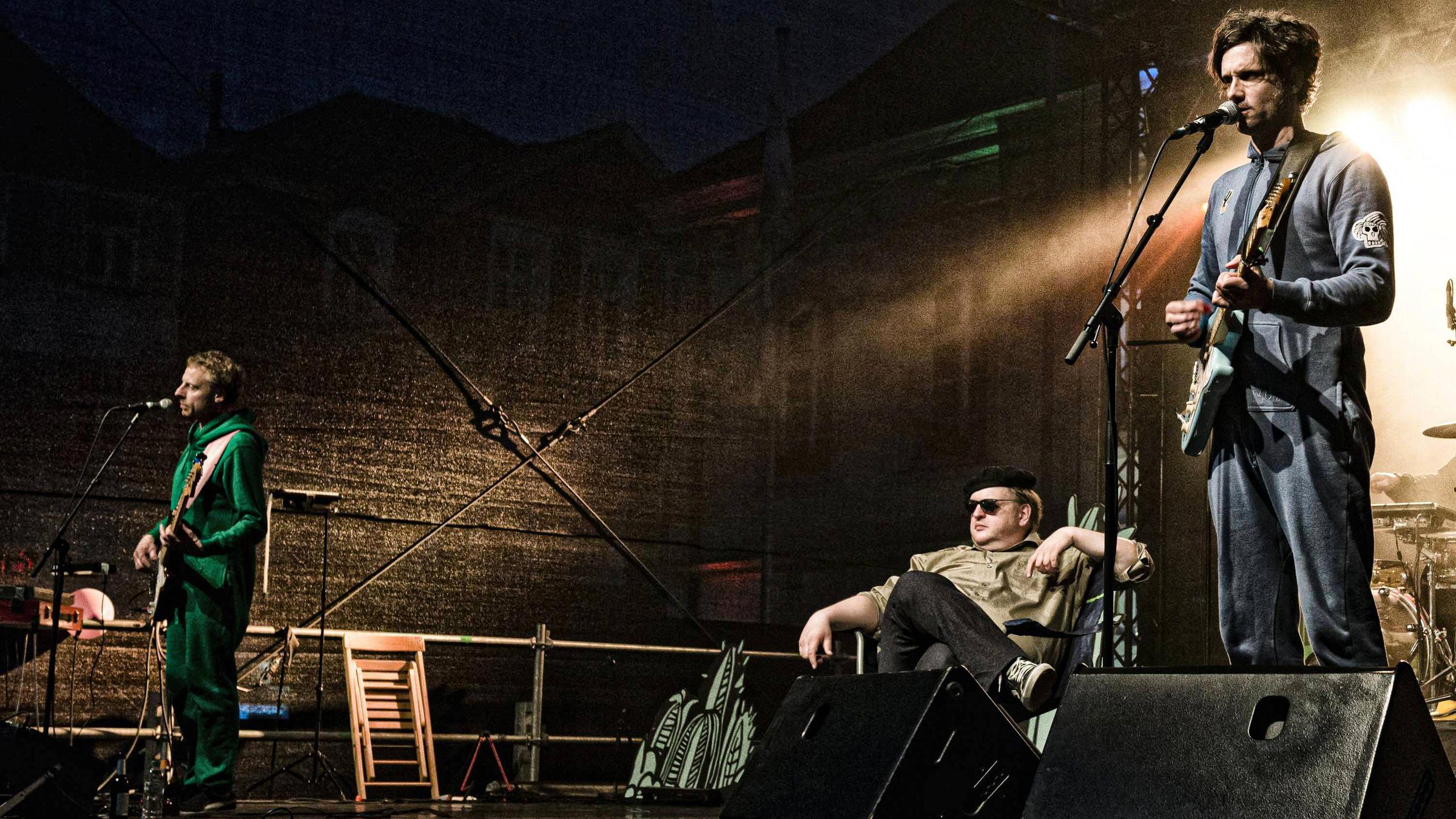 Zwei Männer in Jogginganzügen stehen Gitarre spielend auf einer dunklen Bühne und singen dabei in Mikrofone. Dazwischen sitzt ein dritter Mann mit Sonnenbrille entspannt auf einem Stuhl und schaut gelangweilt ins Publikum.