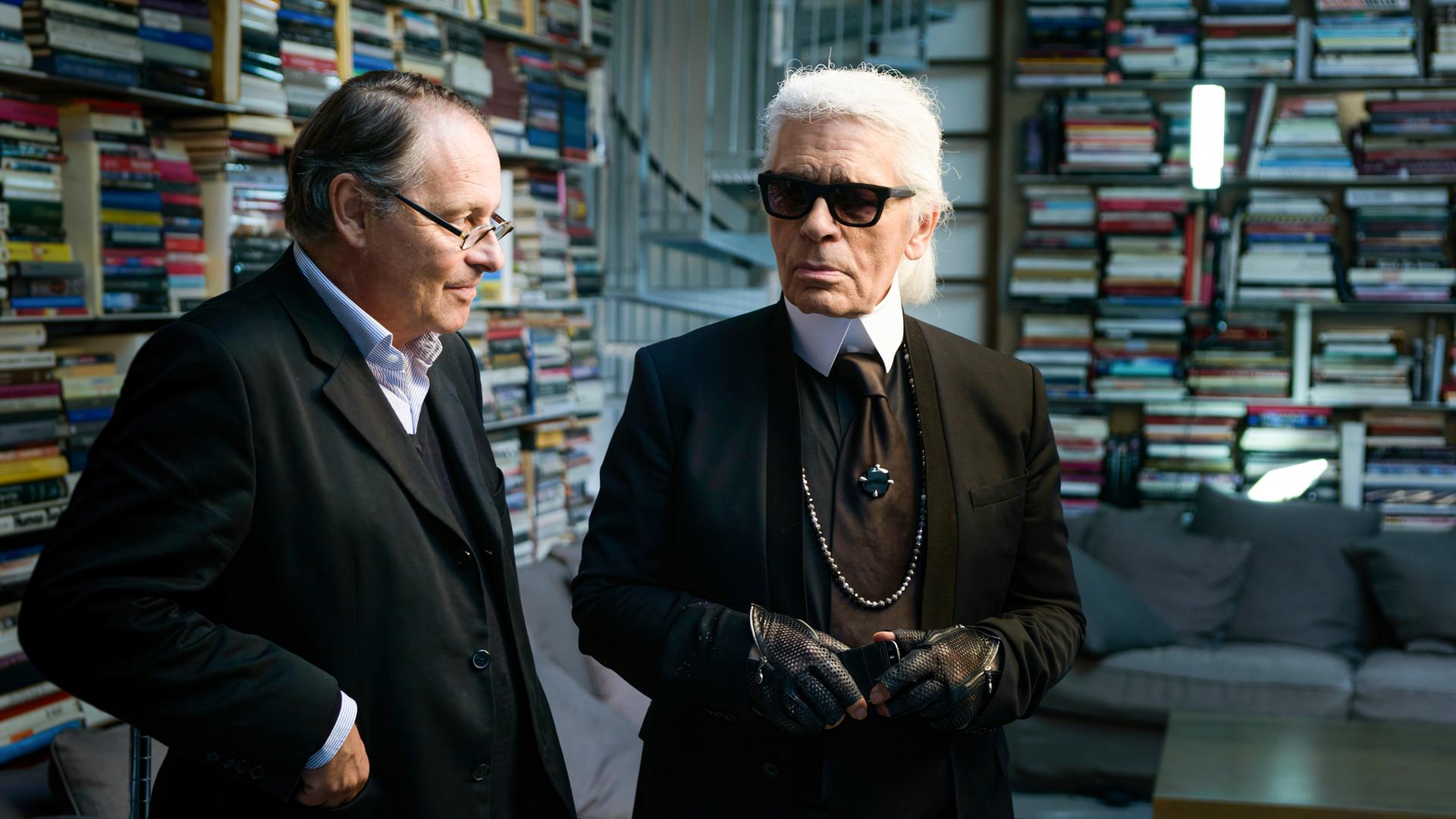 Karl Lagerfeld und Gero von Boehm stehen in einem Raum voller Bücher. Lagerfeld trägt ein schwarzes Outfit mit Krawatte, Sonnenbrille, schwarzen Lederhandschuhen und einer langen Perlenkette. Sein weißes Haar ist streng nach hinten gekämmt. Gero von Boehm, neben ihm, trägt einen dunklen Anzug mit Brille und scheint ihm zuzuhören. Im Hintergrund sind hohe Regale voller bunter Bücher sowie eine Couch mit Kissen zu sehen.
