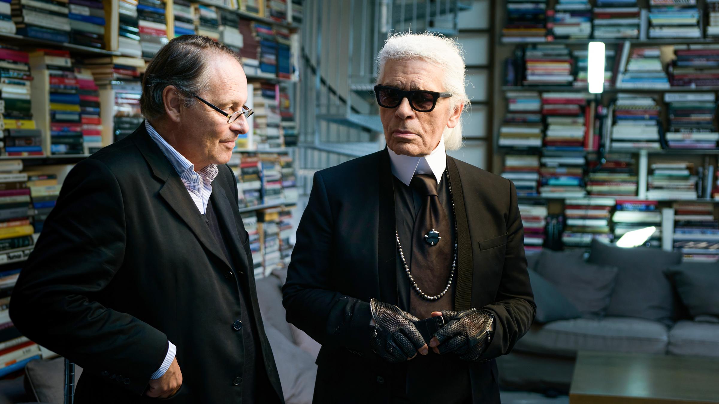 Karl Lagerfeld und Gero von Boehm stehen in einem Raum voller Bücher. Lagerfeld trägt ein schwarzes Outfit mit Krawatte, Sonnenbrille, schwarzen Lederhandschuhen und einer langen Perlenkette. Sein weißes Haar ist streng nach hinten gekämmt. Gero von Boehm, neben ihm, trägt einen dunklen Anzug mit Brille und scheint ihm zuzuhören. Im Hintergrund sind hohe Regale voller bunter Bücher sowie eine Couch mit Kissen zu sehen.