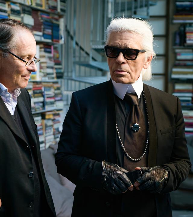 Karl Lagerfeld und Gero von Boehm stehen in einem Raum voller Bücher. Lagerfeld trägt ein schwarzes Outfit mit Krawatte, Sonnenbrille, schwarzen Lederhandschuhen und einer langen Perlenkette. Sein weißes Haar ist streng nach hinten gekämmt. Gero von Boehm, neben ihm, trägt einen dunklen Anzug mit Brille und scheint ihm zuzuhören. Im Hintergrund sind hohe Regale voller bunter Bücher sowie eine Couch mit Kissen zu sehen.