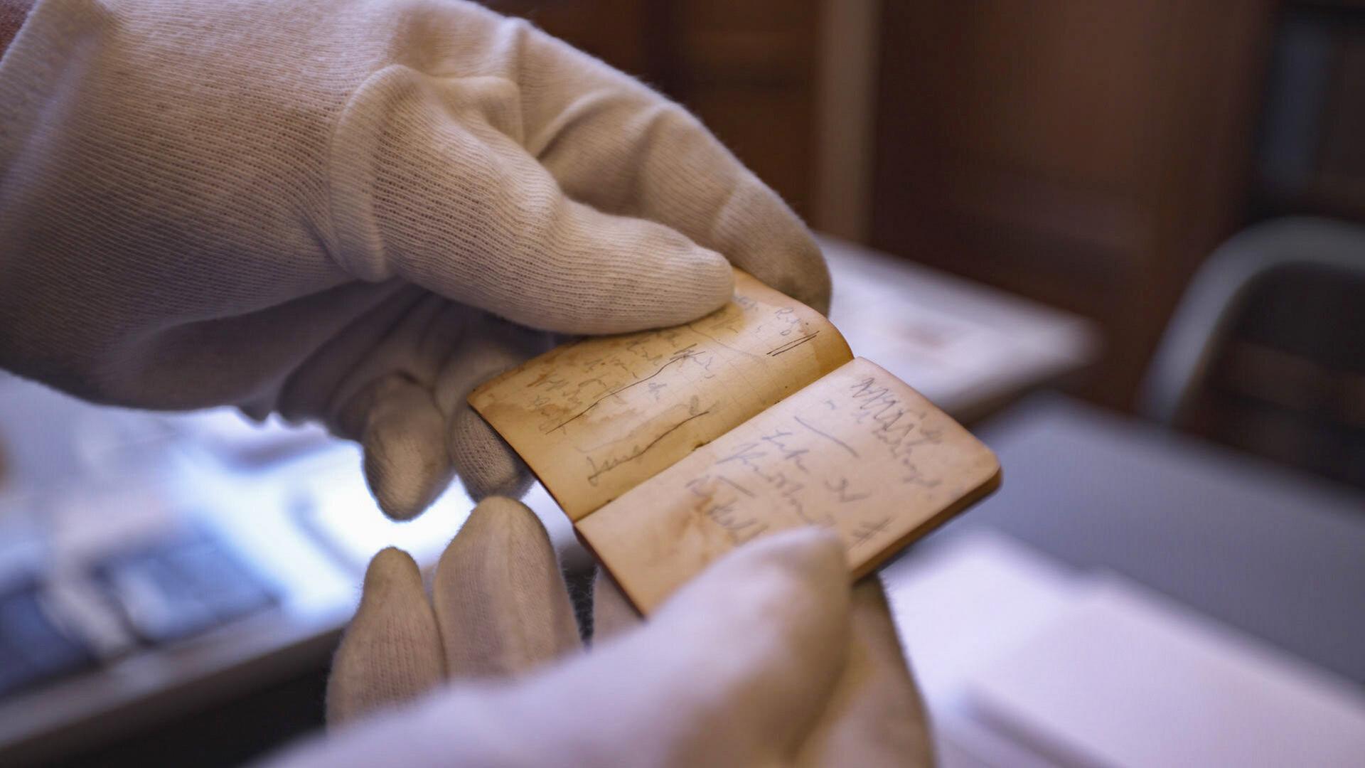 Das Bild zeigt eine Person, die mit zwei Händen ein kleines, aufgeschlagenes Notizbuch hält. Die Person trägt weiße Handschuhe, um das Dokument zu schützen. Das Notizbuch hat einen leicht vergilbten Einband und enthält handschriftliche Notizen, die auf den Seiten sichtbar sind. Die Schrift ist teilweise unleserlich, und die Seiten scheinen im Laufe der Zeit leicht beschädigt oder verfärbt zu sein. Im Hintergrund sind verschwommen einige Möbel und weitere Dokumente zu erkennen, die darauf hindeuten, dass sich die Szene in einem Archiv oder einer Bibliothek befindet.