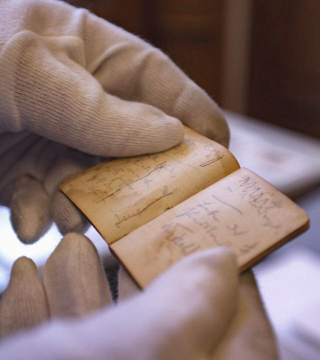 Das Bild zeigt eine Person, die mit zwei Händen ein kleines, aufgeschlagenes Notizbuch hält. Die Person trägt weiße Handschuhe, um das Dokument zu schützen. Das Notizbuch hat einen leicht vergilbten Einband und enthält handschriftliche Notizen, die auf den Seiten sichtbar sind. Die Schrift ist teilweise unleserlich, und die Seiten scheinen im Laufe der Zeit leicht beschädigt oder verfärbt zu sein. Im Hintergrund sind verschwommen einige Möbel und weitere Dokumente zu erkennen, die darauf hindeuten, dass sich die Szene in einem Archiv oder einer Bibliothek befindet.