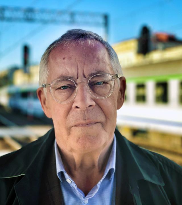 Porträtfoto eines älteren Mannes mit Brille in einem Bahnhof. Im Hintergrund sind Bahngleise und ein Zug zu sehen.