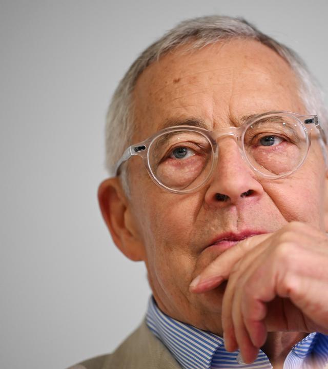 Porträtfoto eines älteren Mannes mit Brille, der nachdenklich eine Hand an den Mund legt.