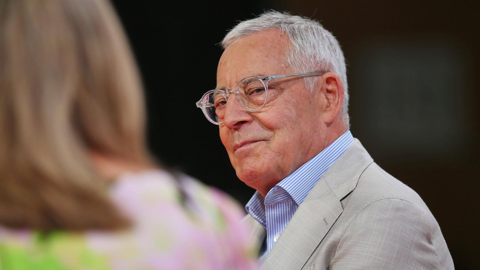 Porträtfoto eines älteren Mannes mit Brille, der sich einer neben ihm sitzenden Frau zuwendet, die nur verschwommen von hinten zu sehen ist.