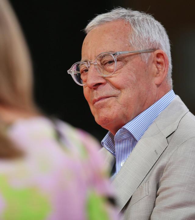 Porträtfoto eines älteren Mannes mit Brille, der sich einer neben ihm sitzenden Frau zuwendet, die nur verschwommen von hinten zu sehen ist.