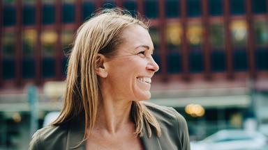 Für die Karriereseiten des ZDF: Eine Frau mit schulterlangen Haaren schaut lächelnd nach rechts, während sie sich draußen vor einer urbanen Kulisse befindet. Der Hintergrund ist weichgezeichnet und vermittelt den Eindruck eines modernen Arbeitsumfelds.