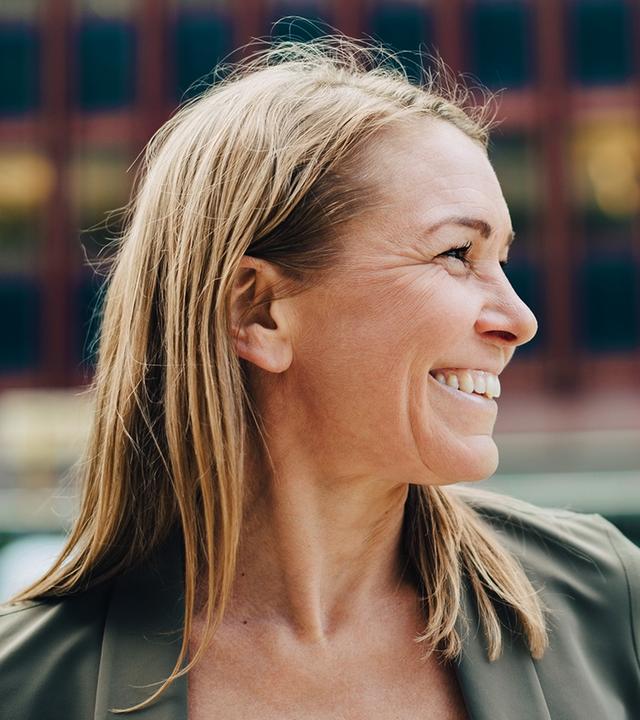 Für die Karriereseiten des ZDF: Eine Frau mit schulterlangen Haaren schaut lächelnd nach rechts, während sie sich draußen vor einer urbanen Kulisse befindet. Der Hintergrund ist weichgezeichnet und vermittelt den Eindruck eines modernen Arbeitsumfelds.
