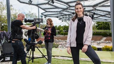 Eine junge Frau steht im Vordergrund und lächelt in die Kamera, während hinter ihr ein Kamerateam mit professioneller Technik arbeitet. Die Szene zeigt eine praktische Arbeitssituation in der Medienproduktion