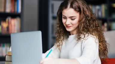 Eine junge Frau sitzt an einem Schreibtisch, nutzt einen Laptop und hält ihre Ergebnisse schriftlich fest. Die Darstellung steht für die Verknüpfung von Studium und praktischer Arbeit im dualen Studienmodell.