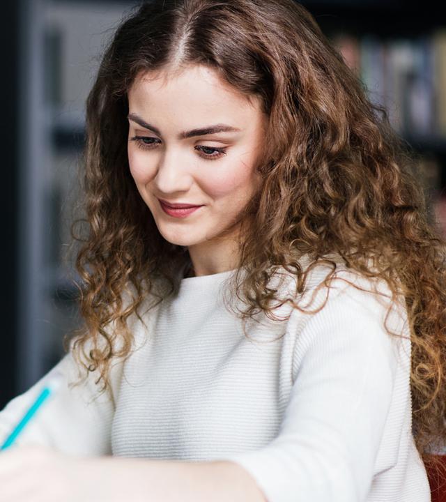 Eine junge Frau sitzt an einem Schreibtisch, nutzt einen Laptop und hält ihre Ergebnisse schriftlich fest. Die Darstellung steht für die Verknüpfung von Studium und praktischer Arbeit im dualen Studienmodell.