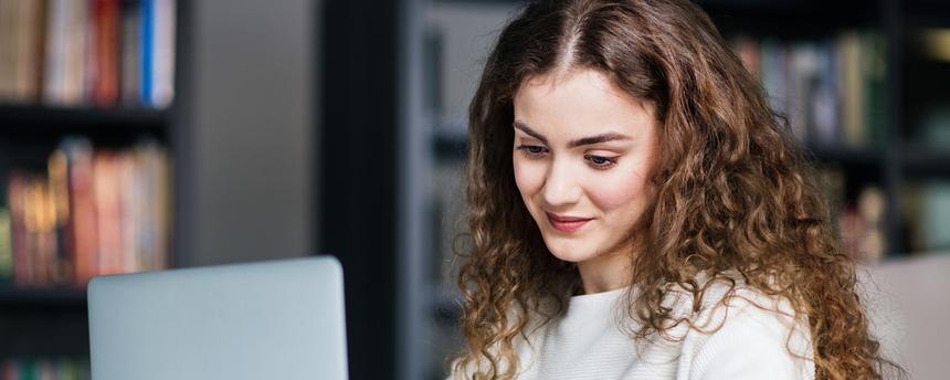 Eine junge Frau sitzt an einem Schreibtisch, nutzt einen Laptop und hält ihre Ergebnisse schriftlich fest. Die Darstellung steht für die Verknüpfung von Studium und praktischer Arbeit im dualen Studienmodell.